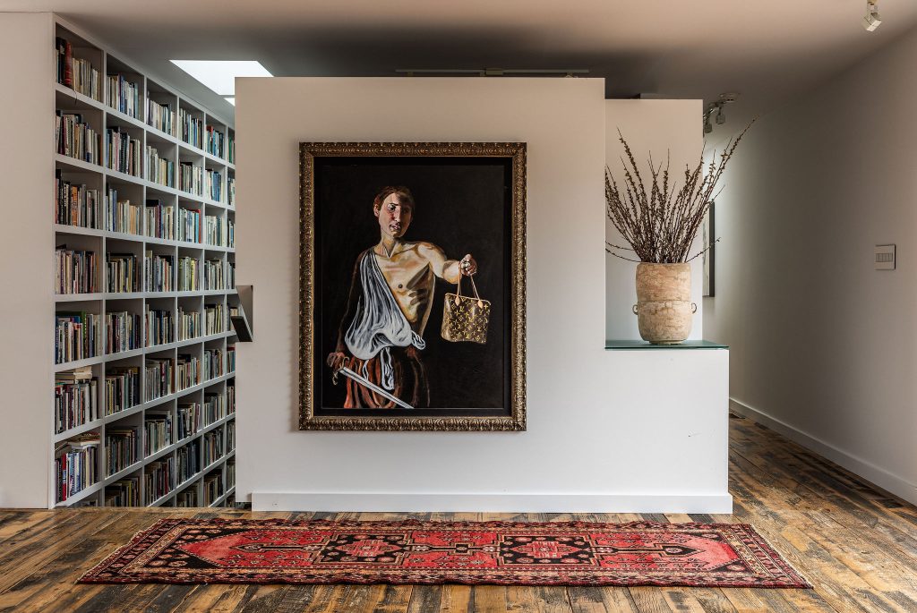 Interior featuring a wall with a large framed painting, a vase with branches, and a colourful rug on wooden floors, next to a built-in bookshelf.