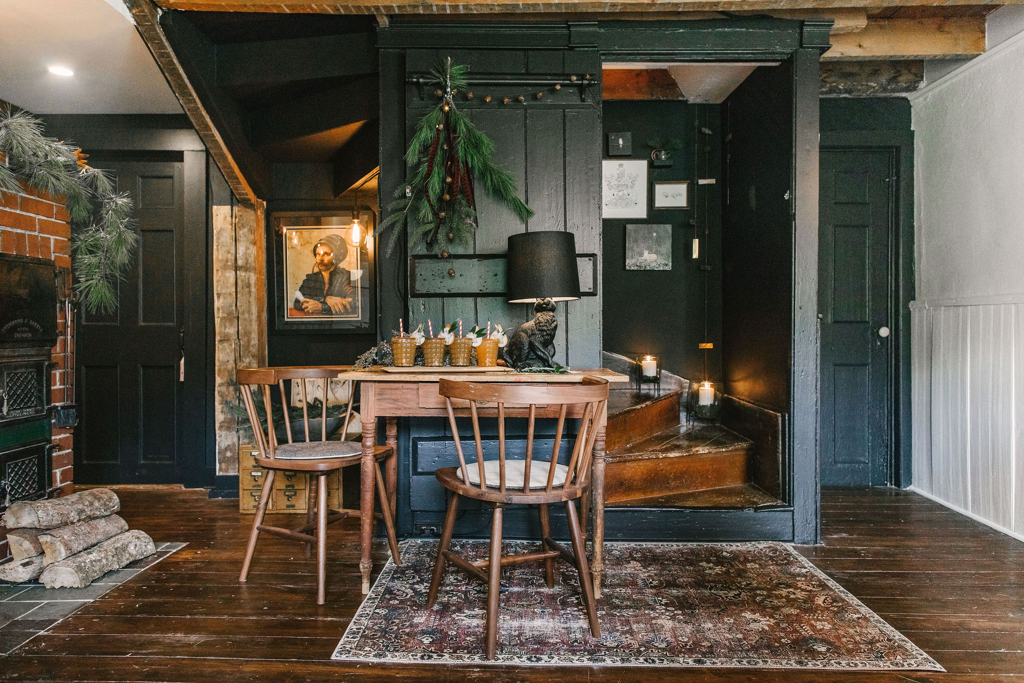 A cozy, rustic dining nook with dark walls, a vintage wooden table, chairs, and a framed portrait. Warm lighting and natural wood elements complement the space, with decorative greenery hanging above the table.