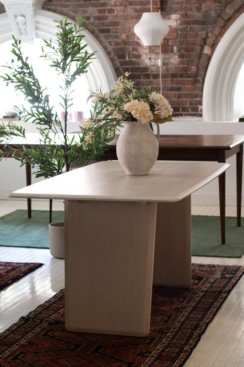 Photograph of a light wood dining table with rounded edges and light wooden plank style legs. Dining Table is on top of a Persian style rug in a retail showroom setting and there is a vase with flowers on the table top.