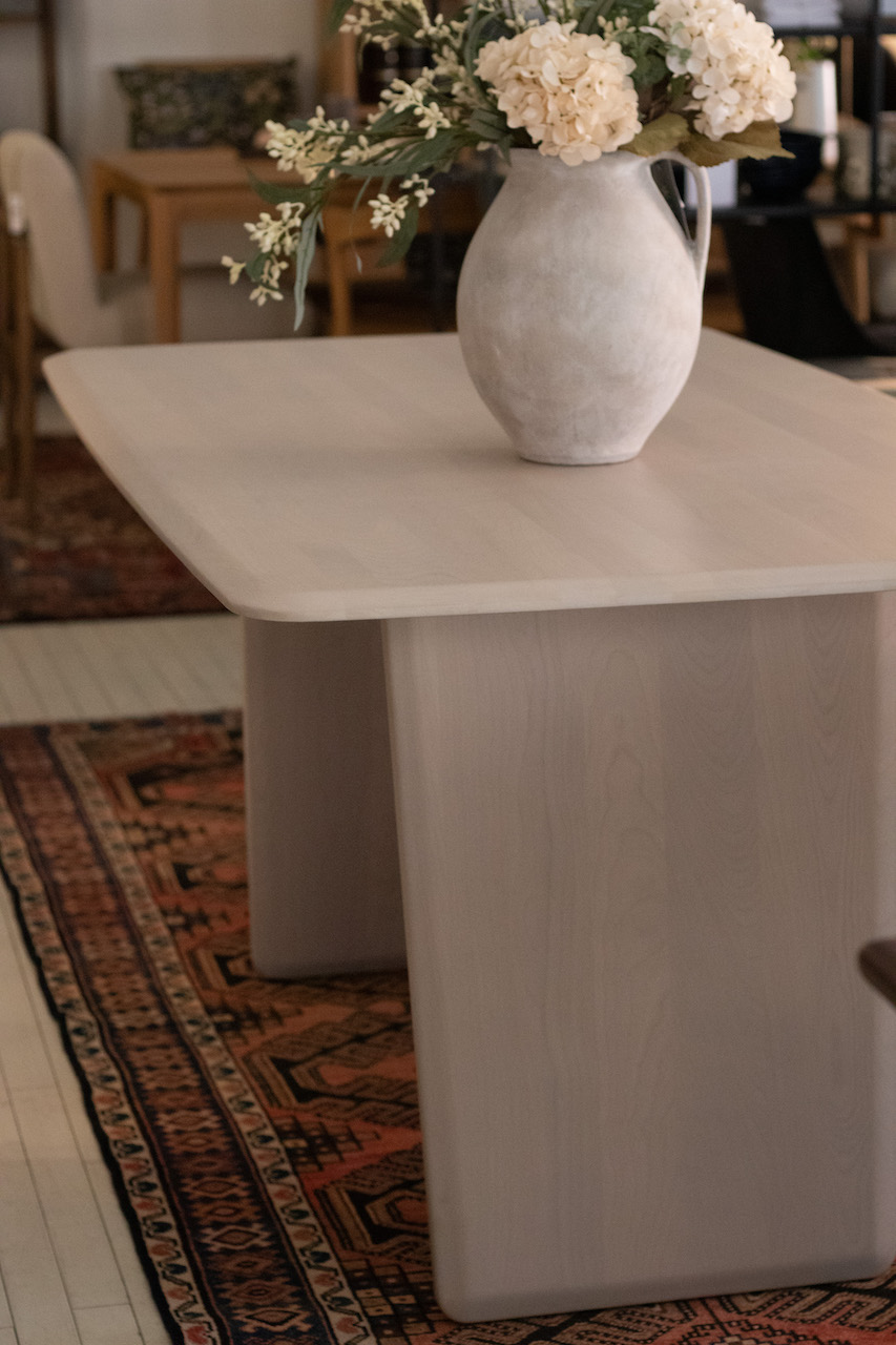 Photograph of a light wood dining table with rounded edges and light wooden plank style legs. Dining Table is on top of a Persian style rug in a retail showroom setting and there is a vase with flowers on the table top.