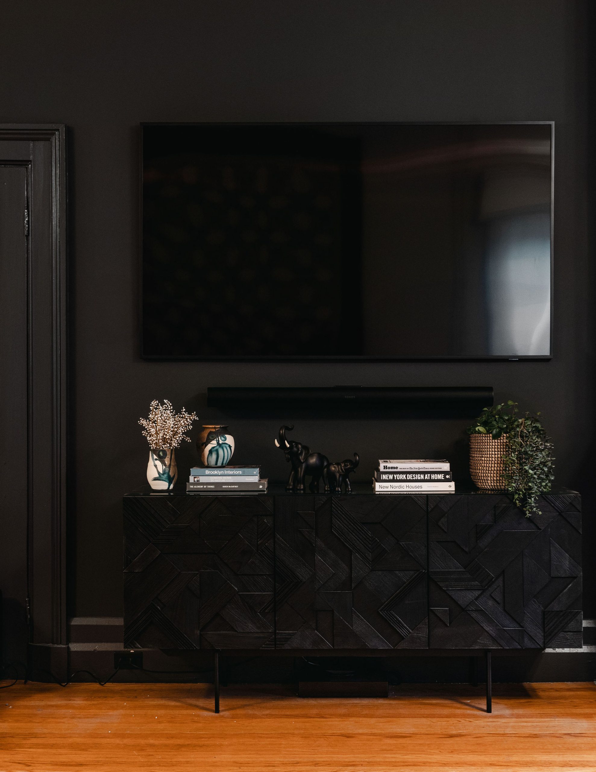 Modern living room setup featuring the Ethnicraft Graphic Dresser in black with geometric carved wood pattern. The dresser is styled with decorative objects including black elephant figurines, a woven planter with trailing greenery, ceramic vases, and stacked coffee table books. A flat-screen TV is mounted above the unit on a dark charcoal wall, with light hardwood floors providing contrast.
