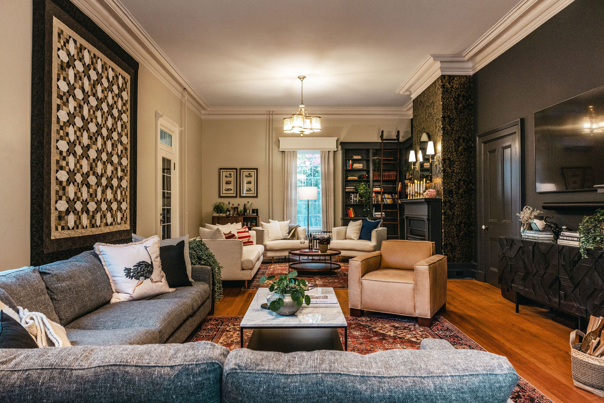 Anderton Roe Great Room 1 A warm, inviting greatroom featuring a rich traditional area rug with red and gold tones, a grey sectional sofa, a pair of cream armchairs, and a tan leather accent chair. The space includes a marble coffee table, dark built-in bookshelves with a rolling ladder, a black fireplace, and a quilted wall hanging. Layered lighting and classic mouldings enhance the architectural charm.