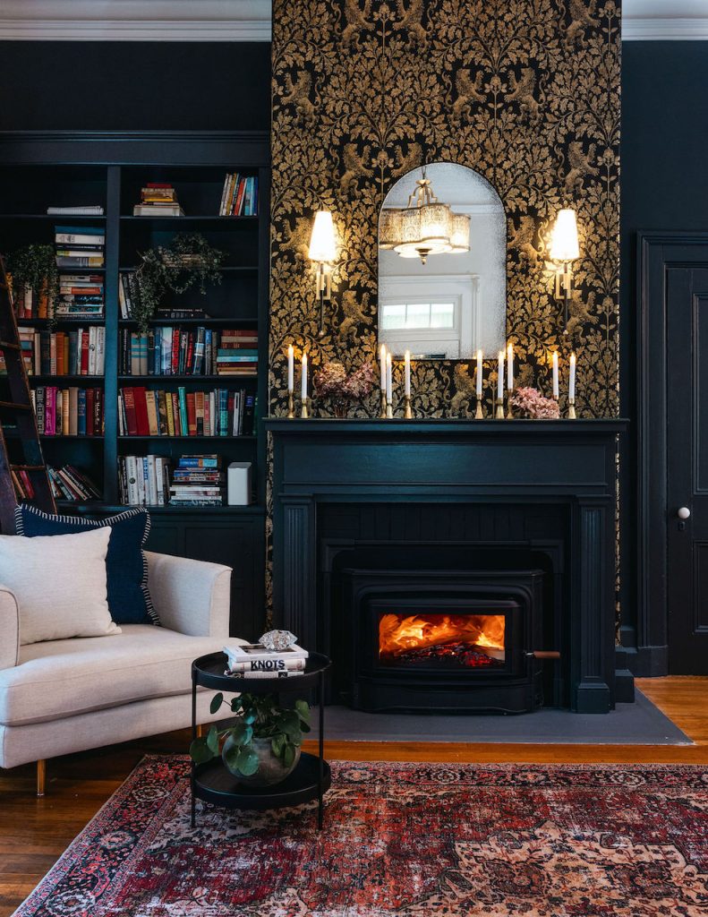 Elegant living room with dark built-in bookshelves and a black fireplace featuring gold Cole & Son Bocabec wallpaper, vintage sconces, and an arched mirror. A cozy fire burns in the wood stove, with a neutral armchair and a round black side table on a red Persian-style rug completing the sophisticated, moody interior design.