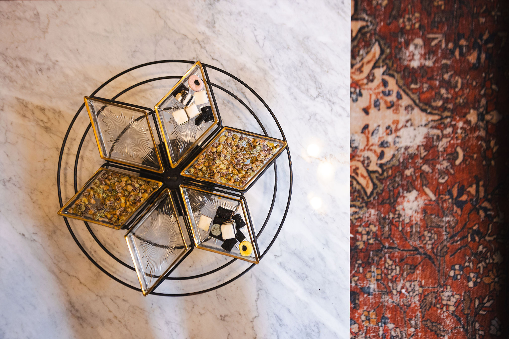 A top-down view of a round serving dish with six star-shaped glass compartments, each trimmed in gold and filled with assorted snacks like pistachios and licorice allsorts. The dish sits on a white marble coffee table with grey veining, adjacent to a richly patterned red and navy vintage-style rug.