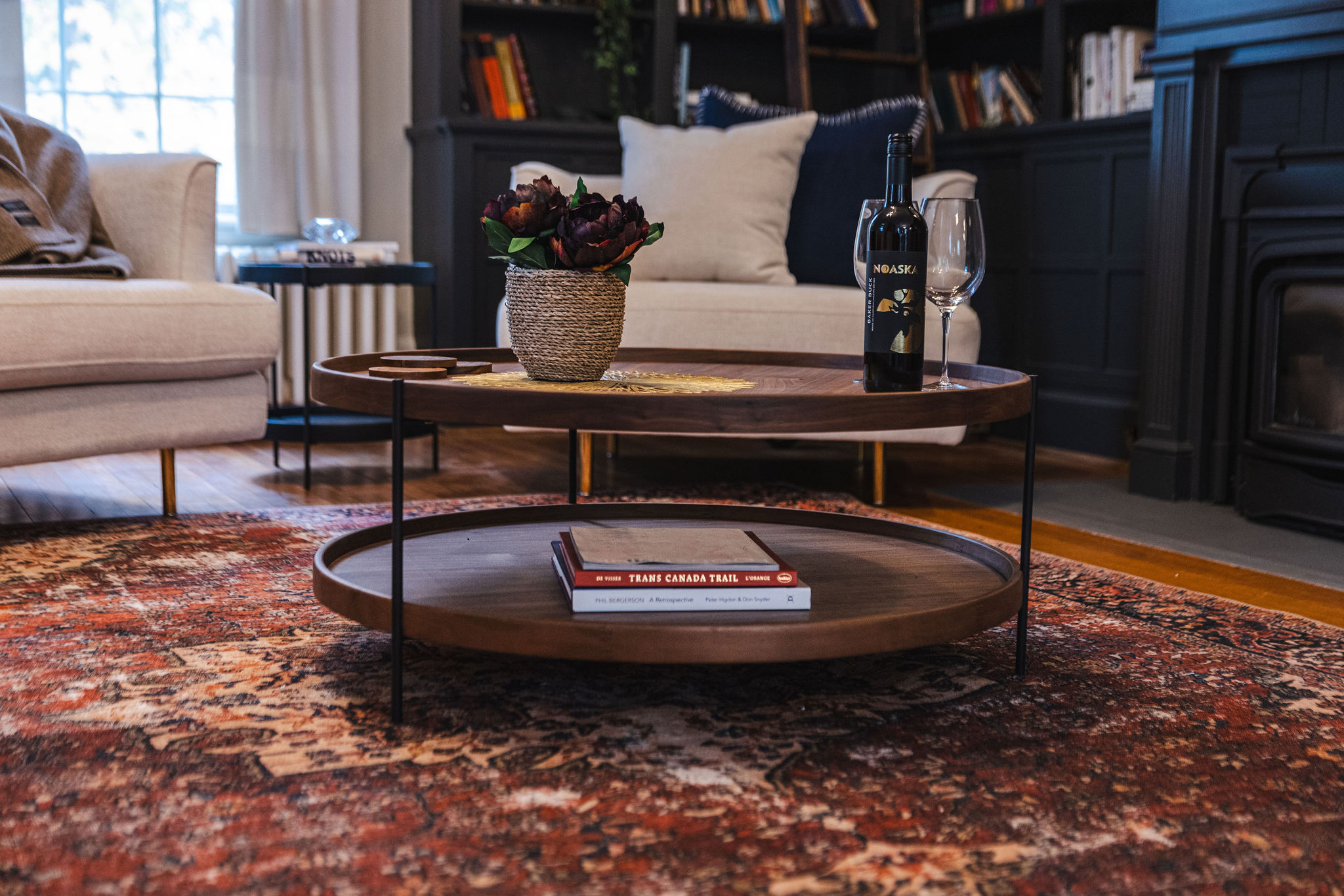 A round walnut coffee table with a lower shelf and slender black metal legs, styled with a woven vase of dark florals, a wine bottle, and glasses on top. The bottom shelf holds a small stack of books, including one titled Trans Canada Trail. The table sits on a richly patterned red vintage rug, surrounded by cream upholstered seating and dark built-in shelving.