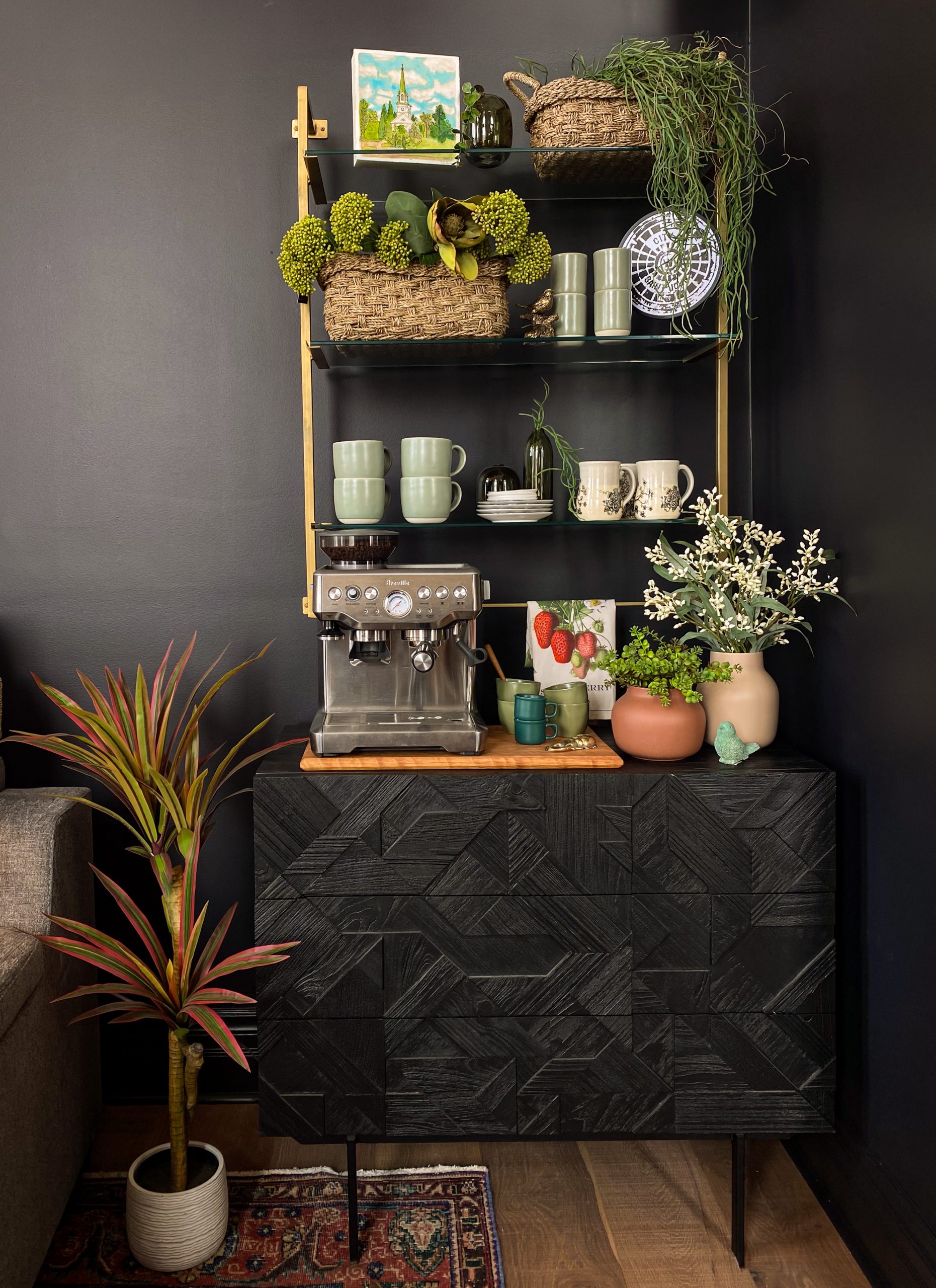 Stylish coffee bar setup featuring the Ethnicraft Graphic Dresser with black geometric wood detailing, topped with a Breville espresso machine and assorted ceramic mugs, vases, and greenery. Above, the Collette wall shelf by Four Hands displays woven baskets, mint green mugs, art, and trailing plants against a dark painted wall. A small vintage-style rug and potted dracaena plant add warmth to the cozy nook.