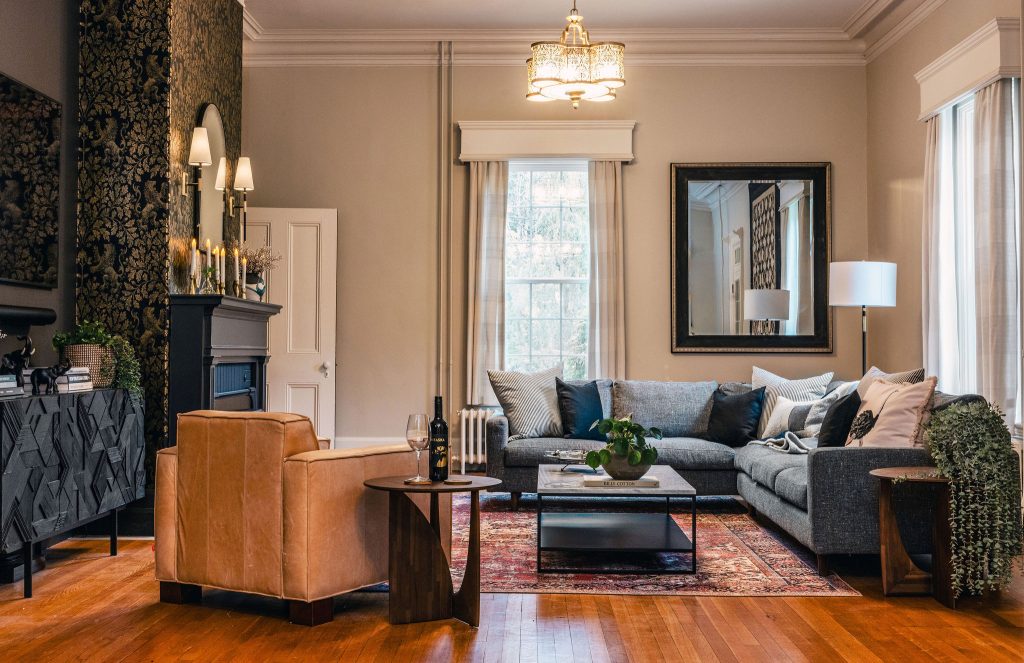 Stylish living room featuring a dark grey sectional sofa with layered neutral and navy pillows, a tan leather armchair, and a vintage-inspired red area rug on warm hardwood floors. A marble-top coffee table sits at the center, flanked by two sculptural wood side tables—one topped with wine and glasses, the other with a trailing plant. A dramatic black fireplace with gold-accented wallpaper and mirror sconces anchors the left wall, while a large mirror and floor lamp reflect light from tall windows dressed in soft drapes. The space exudes a cozy, elegant ambiance with a mix of traditional and contemporary elements.