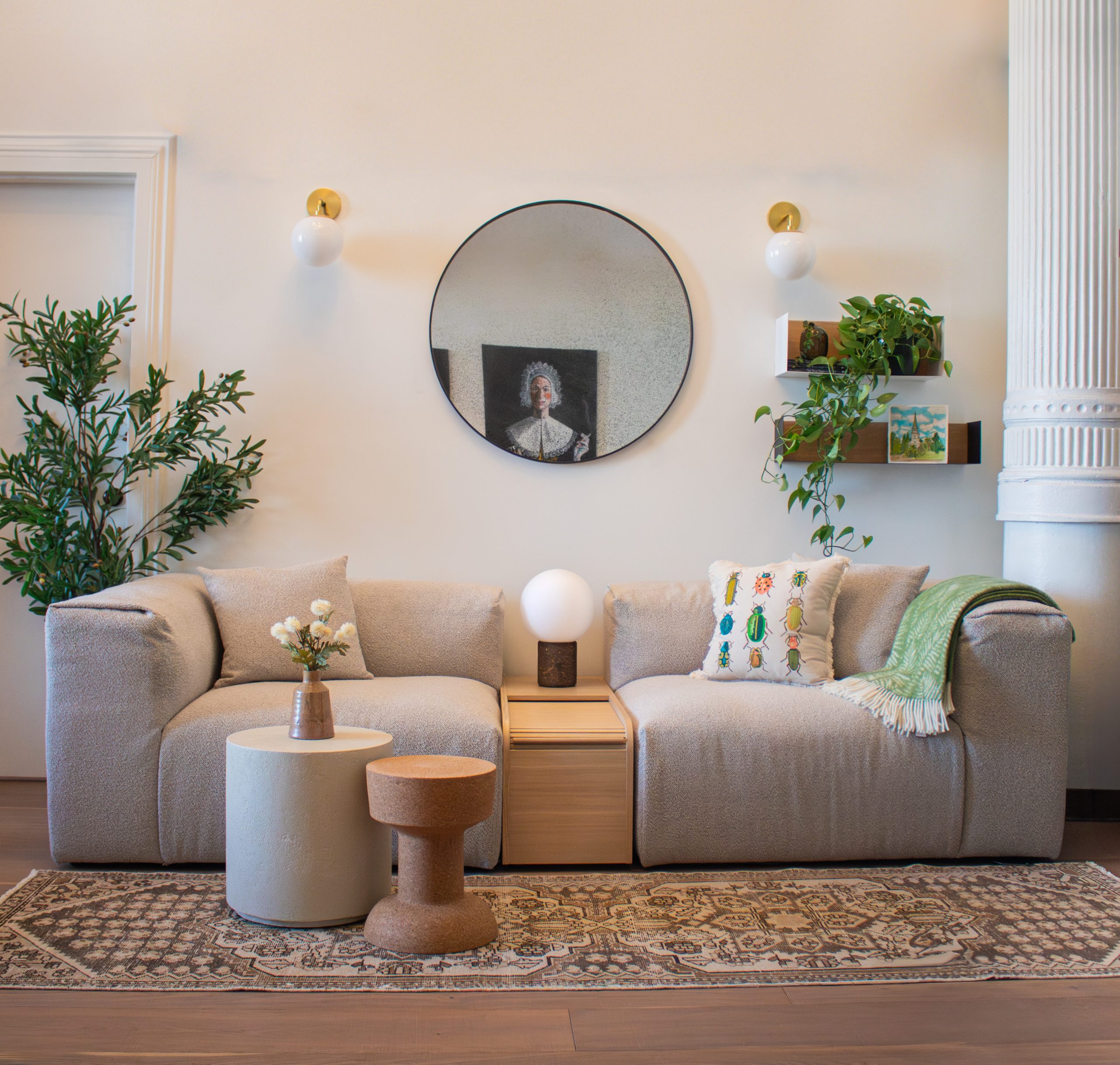 A cozy living room scene featuring the Gus* Modern Next Sofa in a soft, textured fabric with two modular sections flanking the Gus* Modern Ledger Box side table, which holds a round globe lamp. In front of the sofa are two modern accent tables—one cylindrical and one cork. The space is styled with a patterned rug, a round wall mirror reflecting classic portrait art, wall sconces, a hanging shelf with plants and books, and a tall faux olive tree on the left.