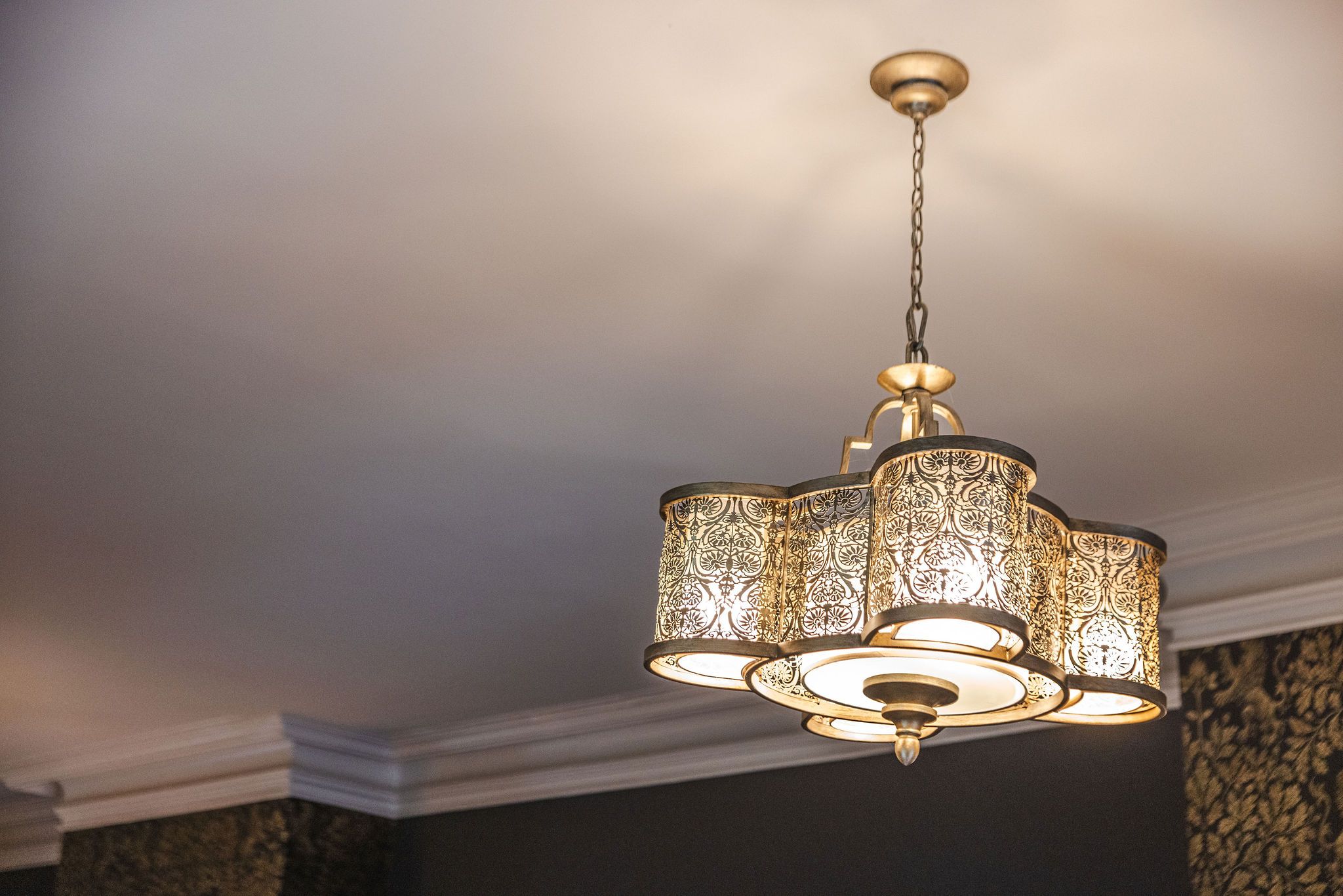 John Catherine Ceiling Light A decorative ceiling light fixture featuring a scalloped drum design with intricate filigree metalwork in a warm gold finish. The chandelier hangs from a short chain and casts a soft, ambient glow, complementing the classic crown moulding and dark, patterned wallpaper of the room.