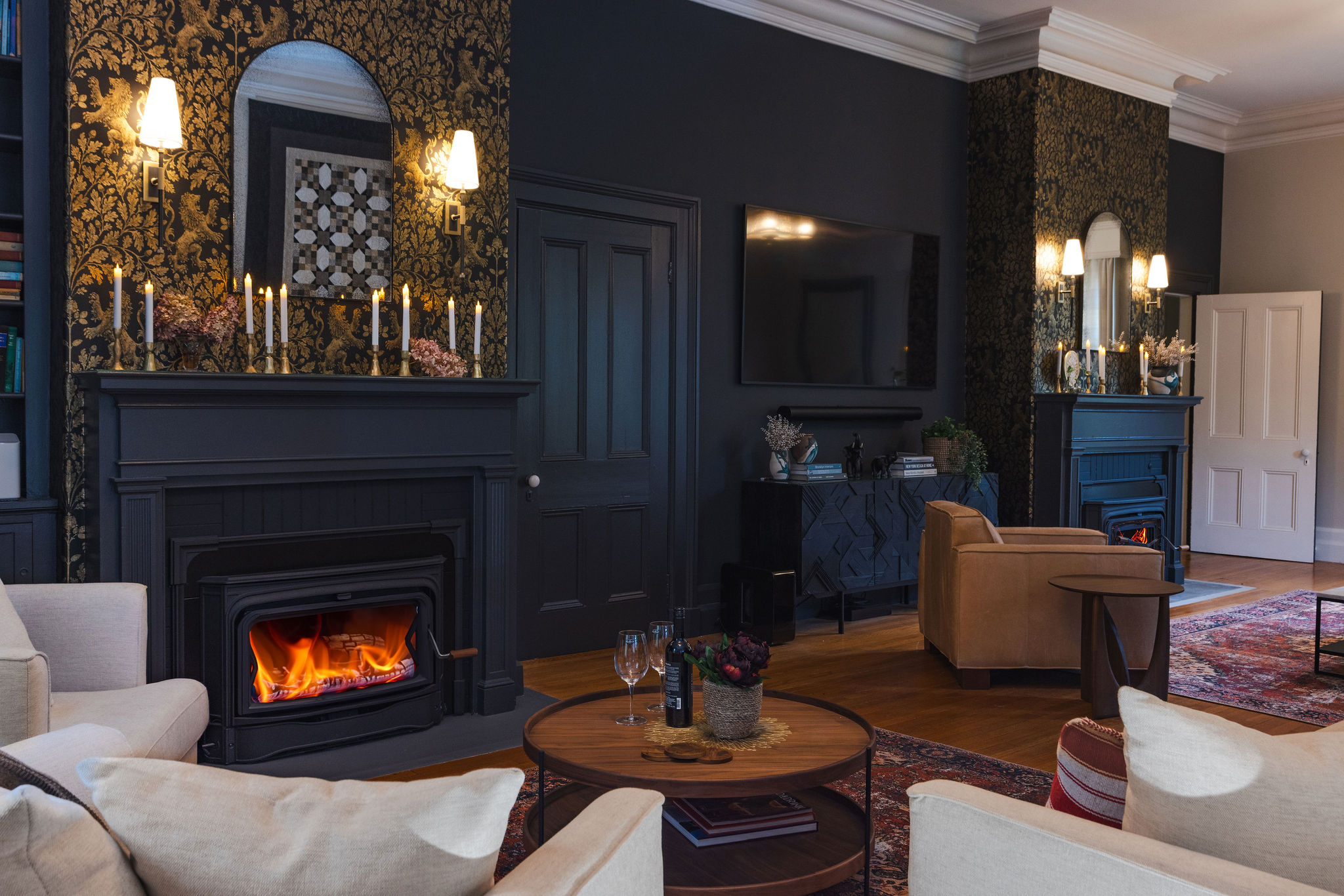 John Catherine Great Room Getaway Fireplaces A sophisticated great room featuring two matching fireplaces adorned with black and gold botanical wallpaper, creating a sense of balance and symmetry. The walls are painted a rich charcoal, and each fireplace mantel is styled with candles, mirrors, and sconces. The room includes a mix of cream and camel-toned seating, layered rugs, and a round wood coffee table. Decorative accents feature mixed metal finishes, books, and plants, creating a warm and curated atmosphere.