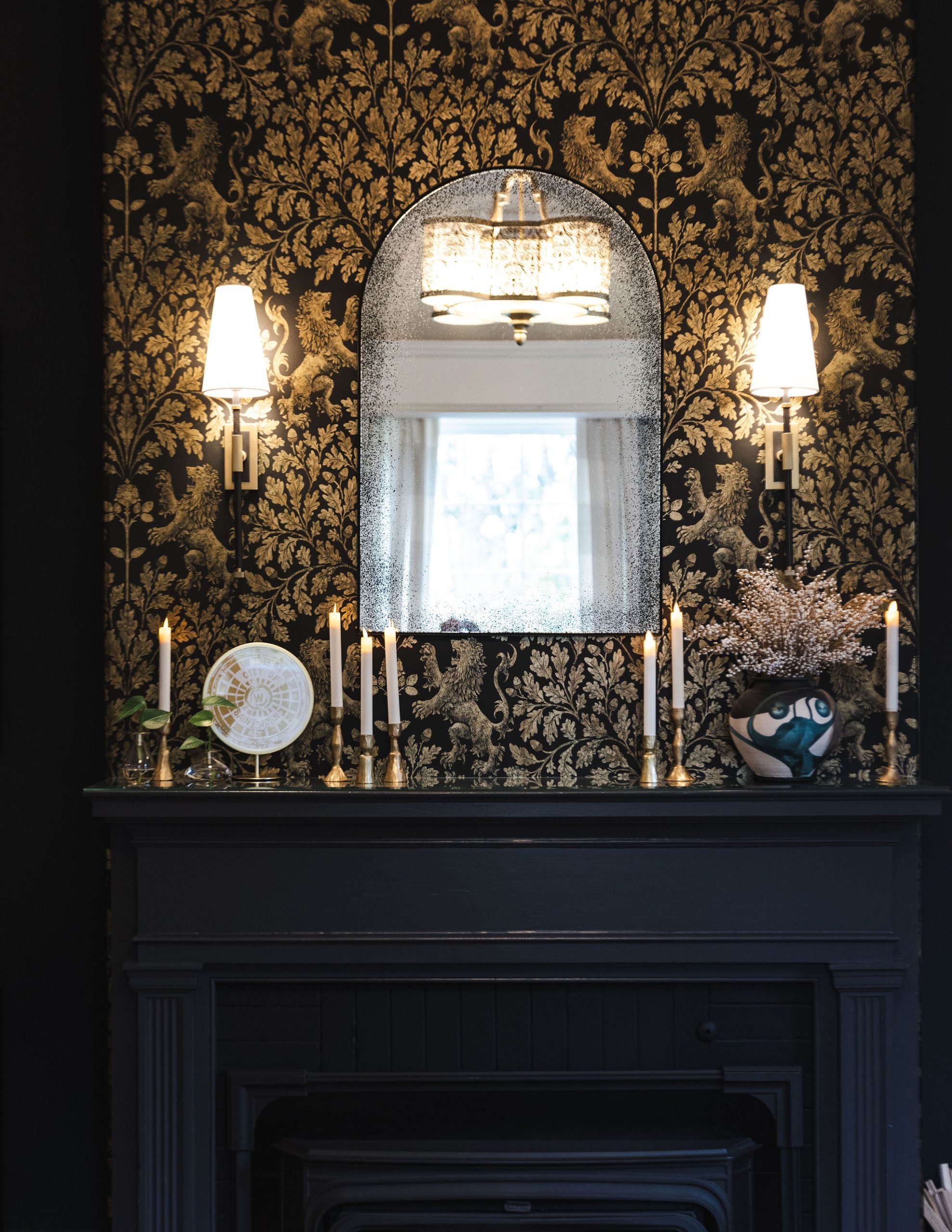 John Catherine Great room Fireplace Wallpaper mirror detail A close-up of a black fireplace mantel set against dramatic black and gold wallpaper featuring a regal lion and vine motif. Above the mantel is an arched antiqued mirror reflecting a chandelier and window. The mantel is styled with brass candlesticks, a decorative plate on a stand, and a sculptural vase with white dried florals. Flanking the mirror are two modern wall sconces with white shades, adding symmetry and ambient light to the richly layered vignette.