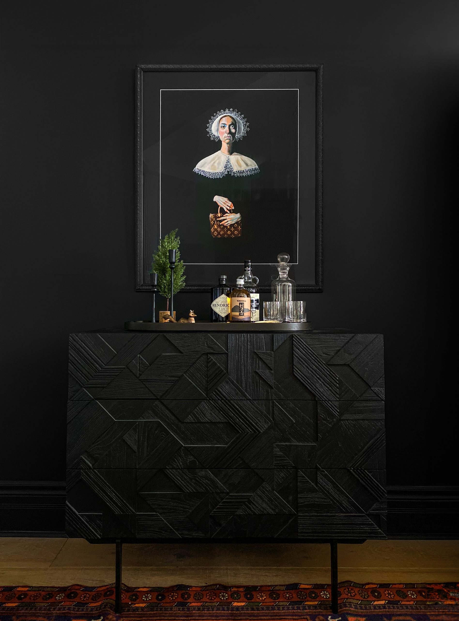 Moody bar vignette featuring the Ethnicraft Graphic Dresser in black with bold geometric wood detailing. Styled as a home bar, the dresser top displays an assortment of liquor bottles, glass decanters, barware, and sculptural bar decor arranged on a round tray. Above, Robert Moore’s ‘Properly Accessorized’ art print in a black frame adds a striking focal point against the matte black wall. A vintage rug peeks out from beneath the dresser, enhancing the rich, dramatic aesthetic.