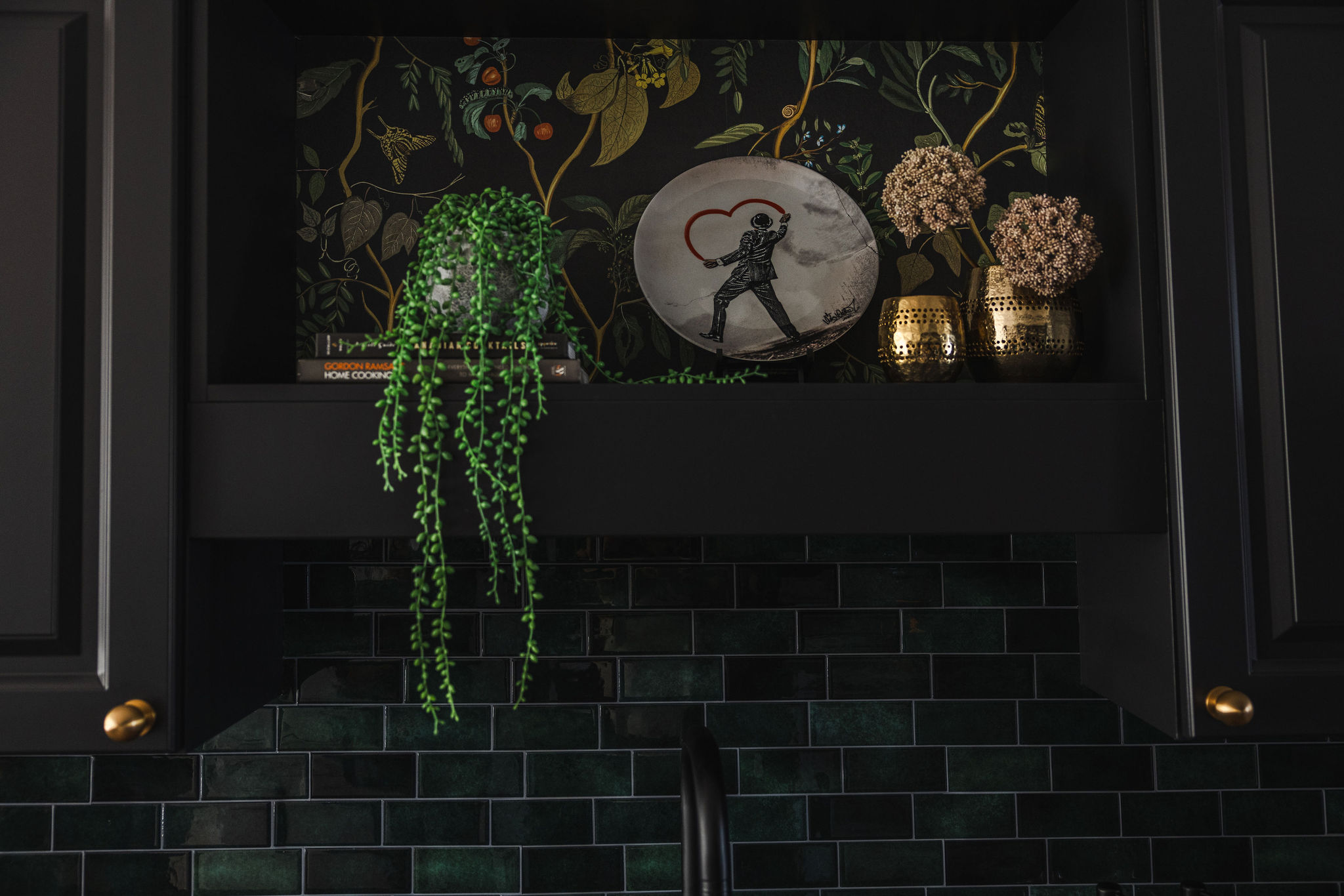 A dark kitchen shelf with a vibrant green trailing plant, art plate featuring a heart motif, brass votives, and floral stems. The shelf is backed by botanical wallpaper and set above glossy deep green subway tiles and matte black cabinetry.