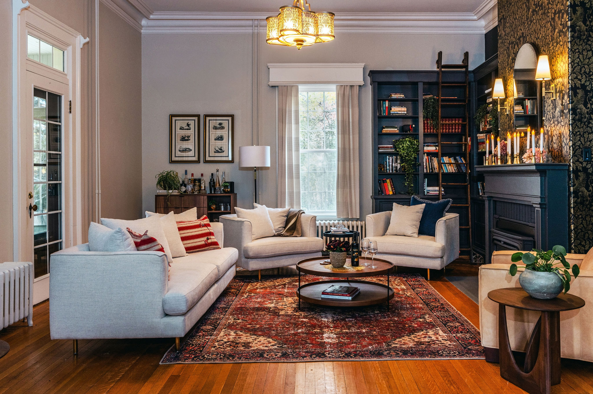 A warm, inviting living room featuring a mix of contemporary and classic design elements. Three light beige upholstered sofas are arranged around a circular coffee table on a rich red Persian-style rug. The backdrop includes built-in navy bookcases with a sliding ladder, a dark-painted fireplace adorned with lit taper candles and a mirror, and wallpaper with a botanical motif. A vintage chandelier hangs overhead, while a mid-century bar cabinet and framed art add charm to the space. Natural light filters through a tall window dressed in soft drapes, and hardwood floors complete the look.