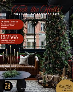Cover of Tuck the Halls! Tuck’s Holiday Lookbook 2025. The scene shows a cozy, festive interior with a tall Christmas tree decorated with lights, ornaments, pinecones, and natural accents. Wrapped presents with neutral kraft paper and ribbon sit beneath the tree. A brown tufted leather sofa holds holiday pillows, including one that reads “Christmas in Saint John.” In front of the sofa is a round black coffee table with evergreen branches and a gold candelabra holding lit candles. The backdrop features large windows adorned with wreaths, revealing a brick building outside. Text highlights inside the lookbook: “Inside Tuck! Our Victorian parlour transformed for the holidays!” and “Shop online and in store – Holiday décor & gifts featured throughout!” Gold and red accents emphasize festive themes, with a note at the bottom right: “100+ pages filled with comfort and joy!