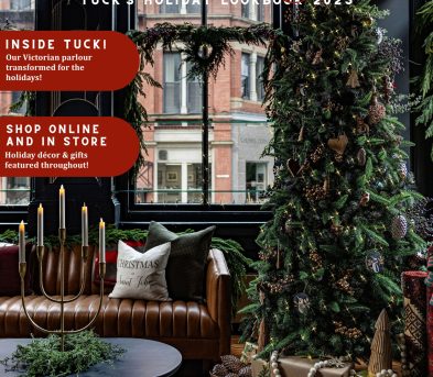 Cover of Tuck the Halls! Tuck’s Holiday Lookbook 2025. The scene shows a cozy, festive interior with a tall Christmas tree decorated with lights, ornaments, pinecones, and natural accents. Wrapped presents with neutral kraft paper and ribbon sit beneath the tree. A brown tufted leather sofa holds holiday pillows, including one that reads “Christmas in Saint John.” In front of the sofa is a round black coffee table with evergreen branches and a gold candelabra holding lit candles. The backdrop features large windows adorned with wreaths, revealing a brick building outside. Text highlights inside the lookbook: “Inside Tuck! Our Victorian parlour transformed for the holidays!” and “Shop online and in store – Holiday décor & gifts featured throughout!” Gold and red accents emphasize festive themes, with a note at the bottom right: “100+ pages filled with comfort and joy!