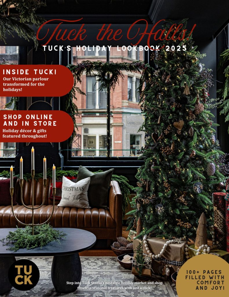 Cover of Tuck the Halls! Tuck’s Holiday Lookbook 2025. The scene shows a cozy, festive interior with a tall Christmas tree decorated with lights, ornaments, pinecones, and natural accents. Wrapped presents with neutral kraft paper and ribbon sit beneath the tree. A brown tufted leather sofa holds holiday pillows, including one that reads “Christmas in Saint John.” In front of the sofa is a round black coffee table with evergreen branches and a gold candelabra holding lit candles. The backdrop features large windows adorned with wreaths, revealing a brick building outside. Text highlights inside the lookbook: “Inside Tuck! Our Victorian parlour transformed for the holidays!” and “Shop online and in store – Holiday décor & gifts featured throughout!” Gold and red accents emphasize festive themes, with a note at the bottom right: “100+ pages filled with comfort and joy!