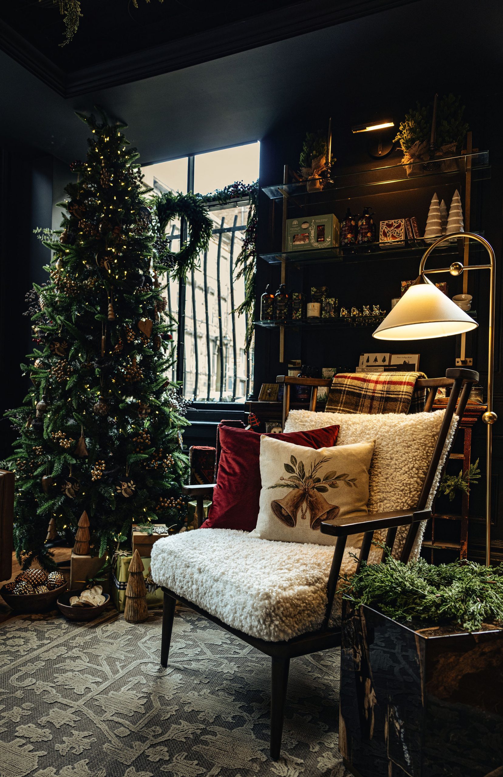 A cozy holiday corner inside Tuck Studio’s Victorian showroom. A Christmas tree decorated with twinkling lights, wooden ornaments, and natural accents stands by a window framed with evergreen garlands. In the foreground, a mid-century inspired chair with a white shearling seat is styled with festive pillows—one deep red velvet and one embroidered with golden holiday bells and greenery. A plaid throw is draped over the chair’s back. To the side, a modern floor lamp casts a warm glow over shelves filled with seasonal décor, gifts, and greenery. Wrapped presents and small decorative trees are arranged at the base of the Christmas tree, creating a warm, nostalgic holiday atmosphere.