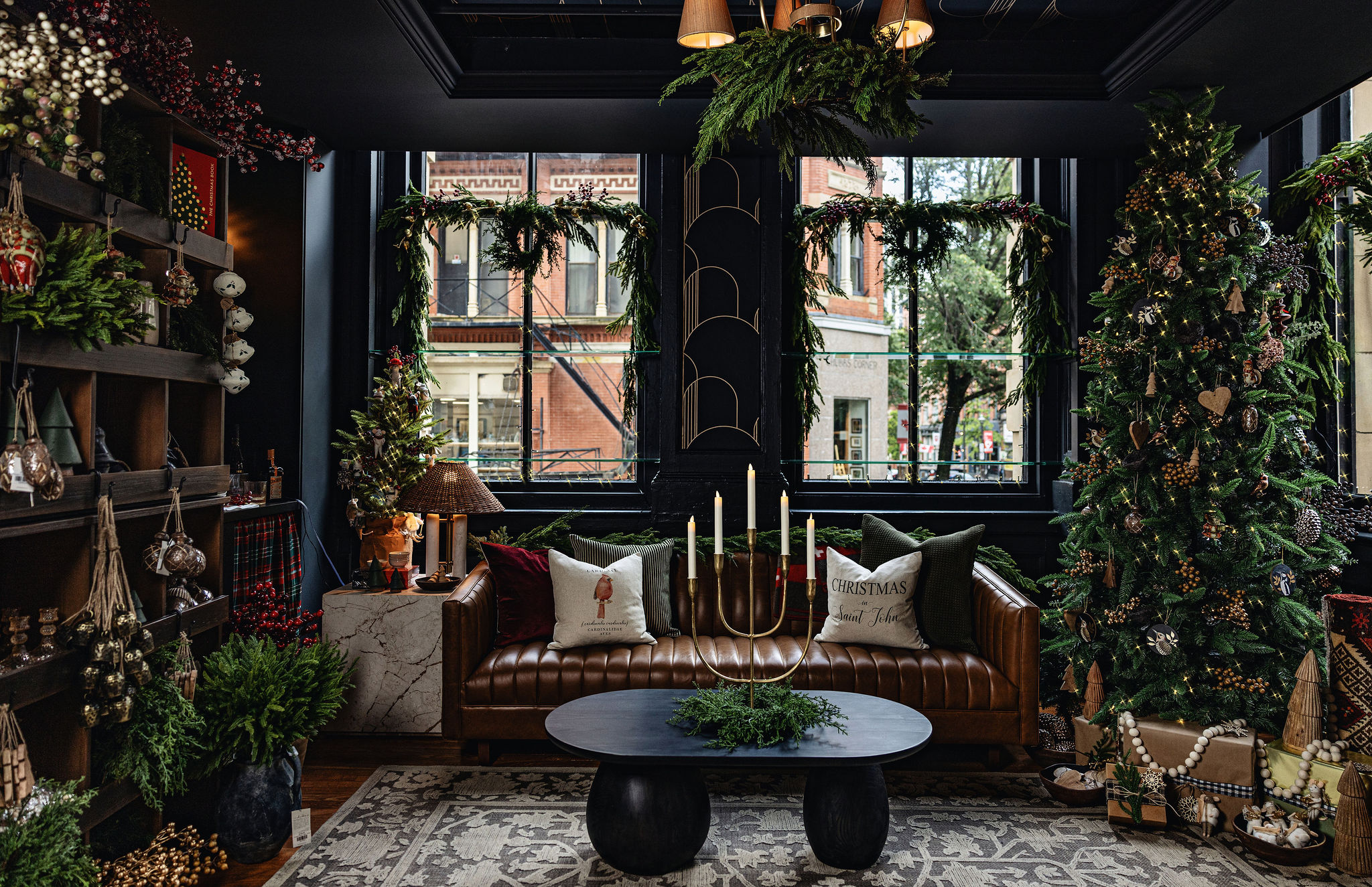 A festive holiday interior inside Tuck Studio’s Victorian showroom. A tall Christmas tree decorated with lights, wooden ornaments, pinecones, and garlands stands to the right, with wrapped gifts beneath. A tufted brown leather Gus* Modern Wallace sofa is styled with holiday pillows, including one embroidered with “Christmas in Saint John” and another featuring a nutcracker. In front, a round black coffee table holds a gold candelabra centerpiece surrounded by greenery. Behind the sofa, large black-framed windows are adorned with evergreen garlands and wreaths, offering a view of historic brick buildings in Uptown Saint John. To the left, wooden shelves display festive décor: hanging bells, ornaments, greenery, and seasonal accents. The overall atmosphere is cozy, nostalgic, and elegant with Victorian holiday charm.