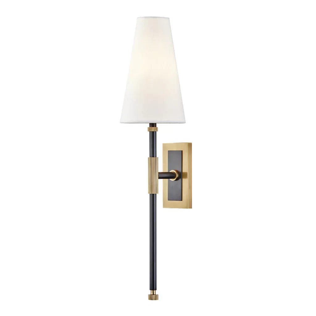 A modern wall sconce featuring a tapered white fabric shade atop a slender black metal rod with brass accents. The fixture is mounted on a rectangular brass and black backplate, combining classic and contemporary elements in a sleek, vertical design.