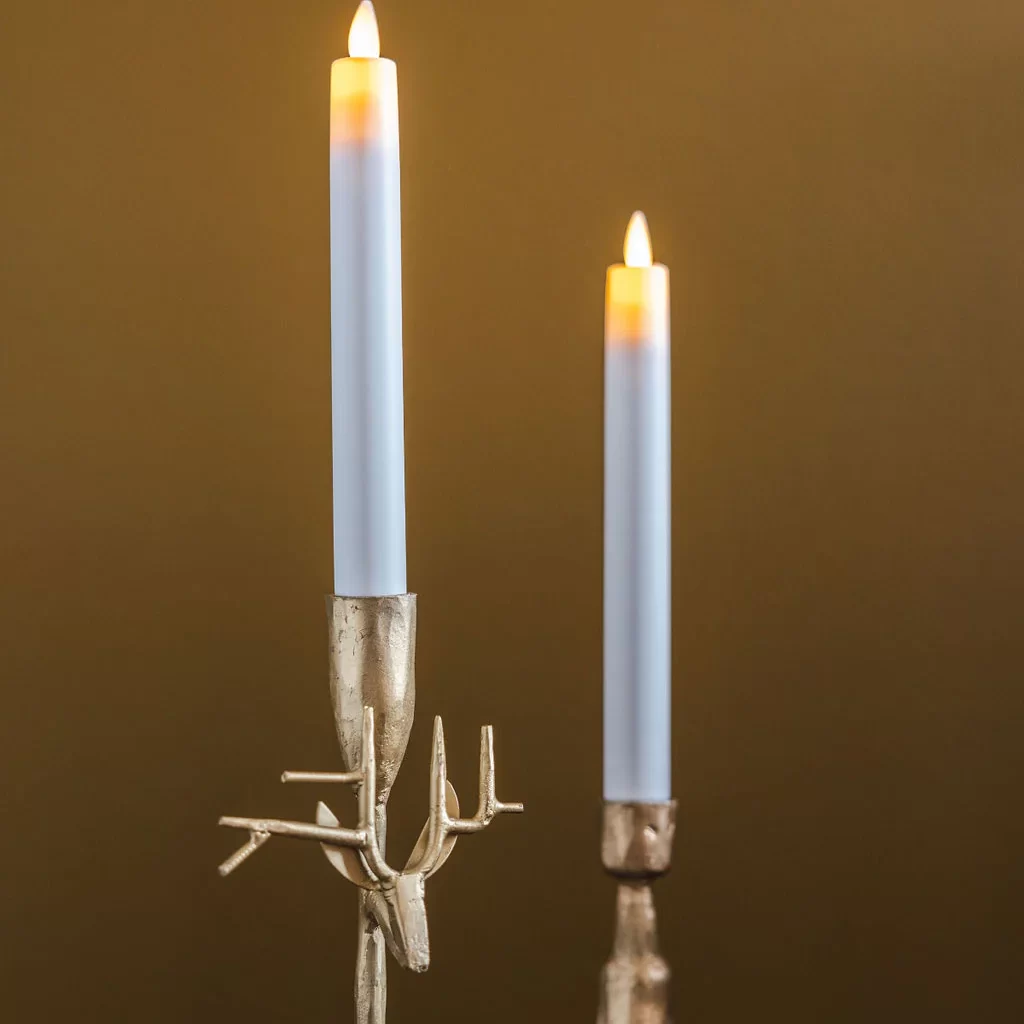Two battery-operated taper candles with warm glowing LED “flames,” set in sculptural gold holders. One holder features a stylized deer head with antlers, while the other has a textured, organic shape. The candles stand on a dark reflective surface against a warm mustard-toned background.